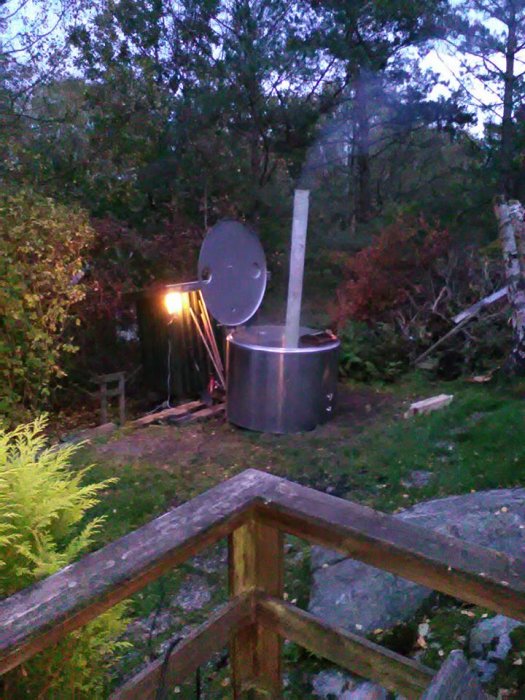 Däck med rostfri mjölktank omvandlad till badtunna med aluminiumkamin, rök stiger upp, omgiven av natur.