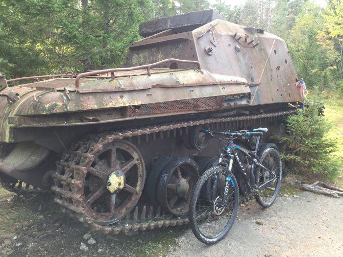 En mountainbike parkerad framför en gammal rostig stridsvagn i en skogsmiljö.