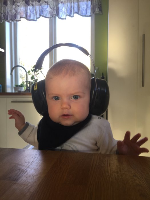 Bebis med stora hörlurar sitter vid bord, ser in i kameran, kök i bakgrunden.