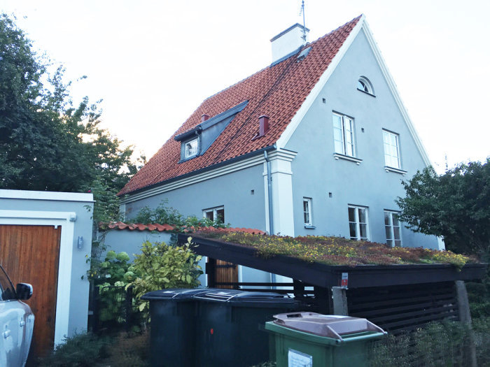 Cykelställ med grönt tak framför hus med röd takpannor och vit putsad fasad.