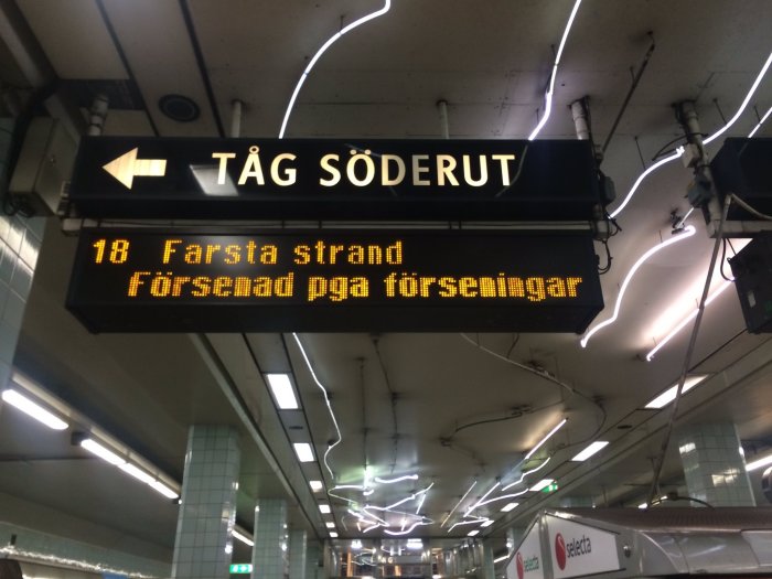 Tågstationsskylt med text "TÅG SÖDERUT 18 Farsta strand Försenad pga förseningar".