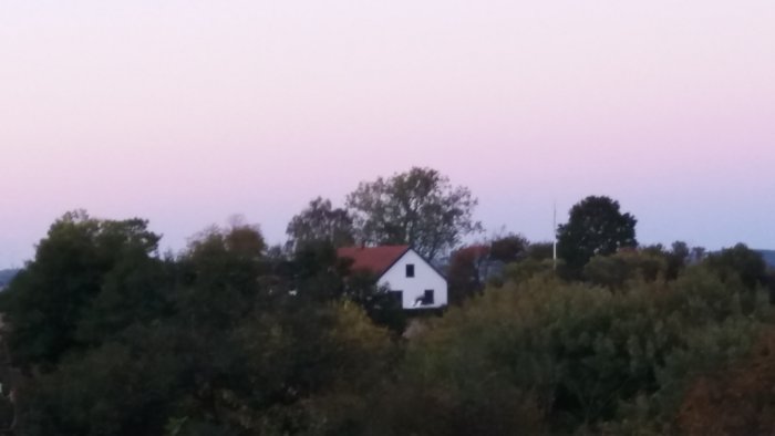 Skymning med mjuka rosa himmelstoner, vy över trädtoppar med en vit villa med rött tak i bakgrunden.
