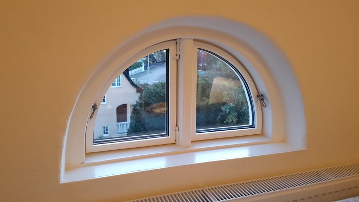 Renoverat halvmåneformat vindsvåningsfönster med vita karmar och spröjs mot en gul vägg.