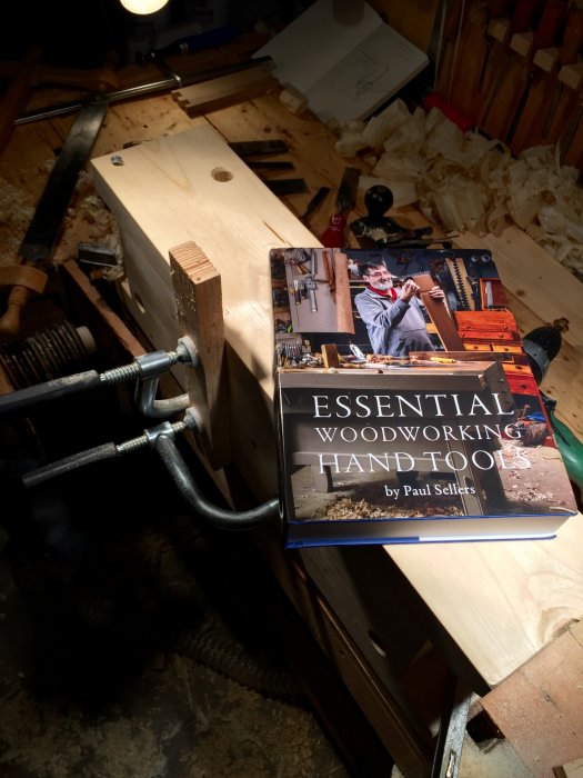 Bok om träbearbetning, "Essential Woodworking Hand Tools" av Paul Sellers, ligger på en snickarbänk omgiven av verktyg och träspån.