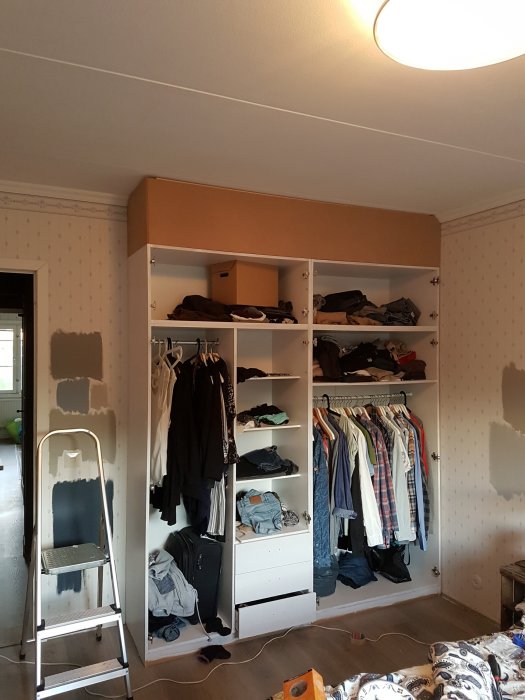 Förvandlad fristående garderob i ett sovrum under renovering, med öppna fack och kläder, intill en stege.