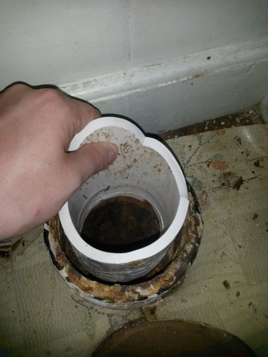 Hand som håller en vit muff över ett rostigt gjutjärnsrör i golv, omringad av gammal silikon och smuts.