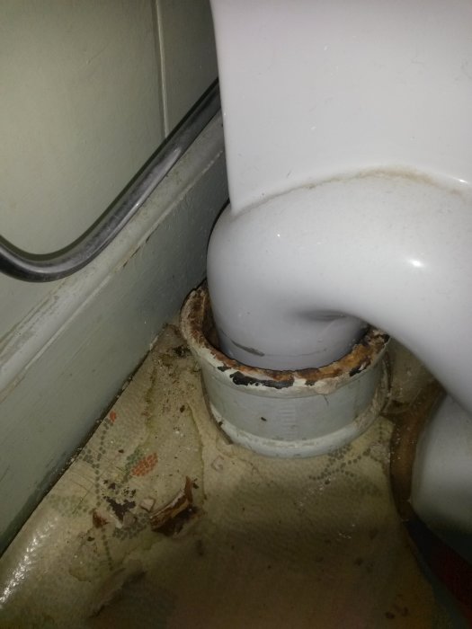 Glipan mellan en vit toalettstol och röranslutningen på golvet, med tecken på läckage och korrosion.
