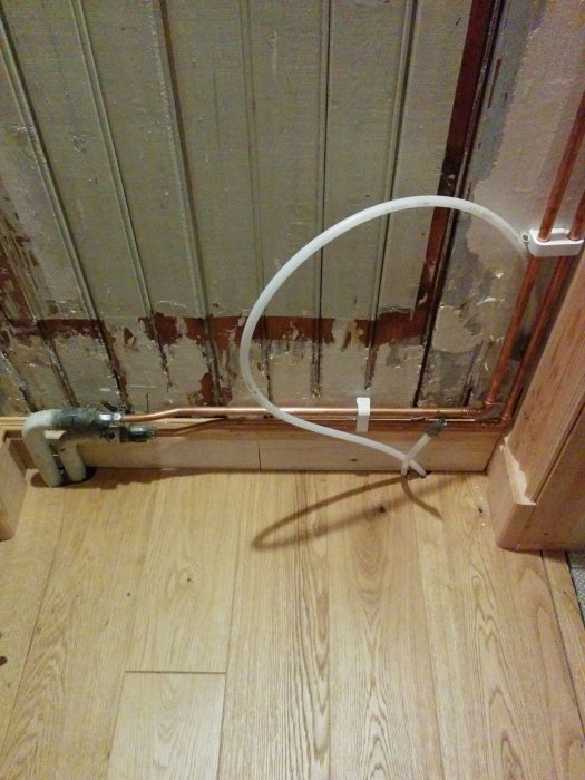 Rörinstallation i en utbyggd hall med synligt rörarbete och kabel under klinkergolv vid en vägg.