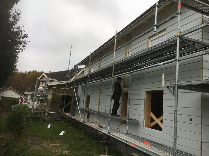 Arbetare på byggställning framför hus med delvis monterad träpanel.