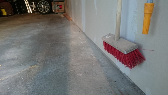 Röd sopborste lutad mot en ljus vägg i ett garage, med del av en bil och hyllor i bakgrunden