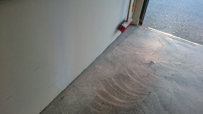 Nyputsad vit vägg och betonggolv med röd kvast i hörnet vid en dörröppning.