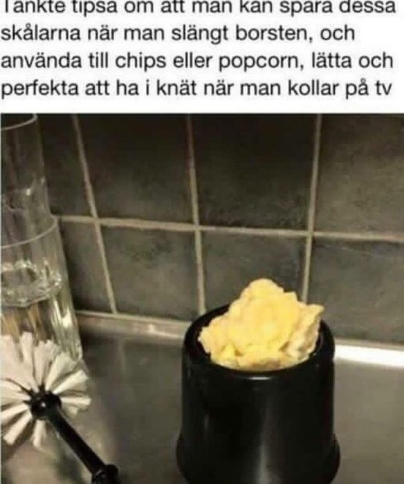 Svart toalettborstkärl återanvänt som snacks-skål fyllt med chips står på köksbänk.