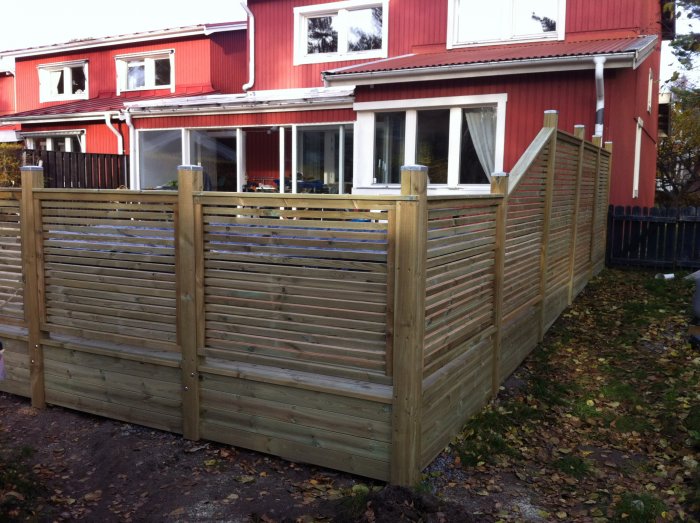 Trädgårdsstaket av trä med stolpar fästa i ramen, beläget framför ett rött hus.