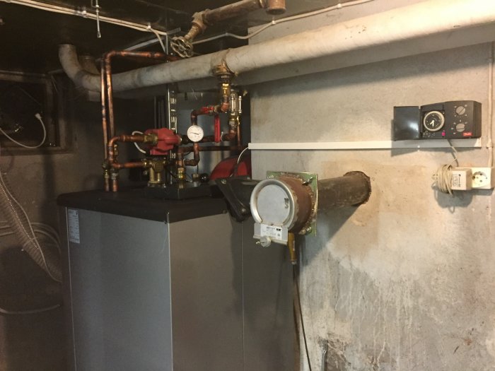 Källare med värmepanna, rörinstallationer, termostat och eluttag på eternitvägg.