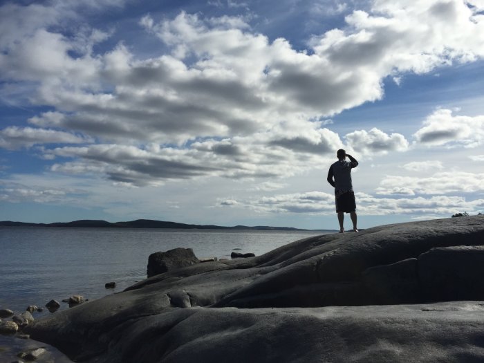 Person står på klippan vid havet i Höga Kusten med molnig himmel under midnatt på midsommar.
