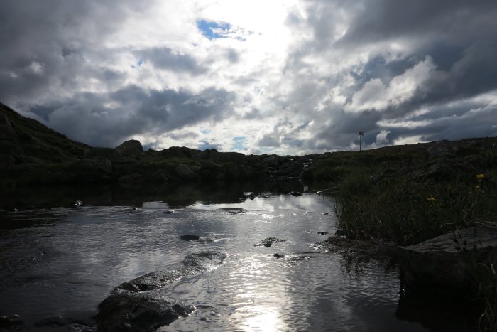 Dramatisk himmel reflekterad i vatten med klippor och vegetation, molnigt väder.