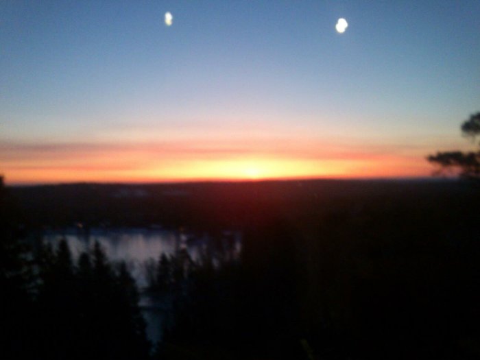 Solnedgång med mjuka röda toner över en skog och sjö, med diffust ljus och suddig bakgrund.