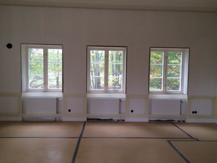 Ett rum med golv till tak-fönster och tejpade markeringar för spegelpanel längs väggen ovanför radiatorer.