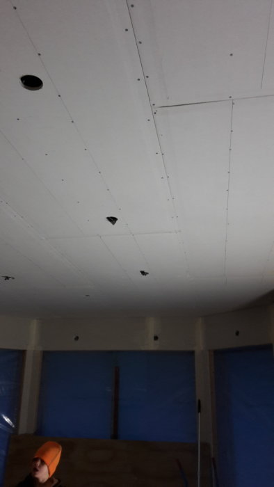 Inre takkonstruktion med gipsskivor och synliga skruvar, oavslutad installation, person i orange mössa tittar upp.