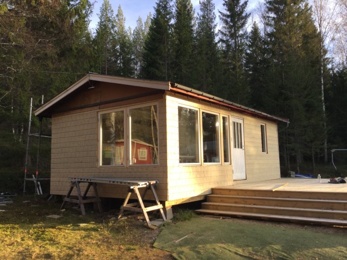 Ett litet hus under renovering med nybyggd stor trädäck i förgrunden, omgivet av skog.