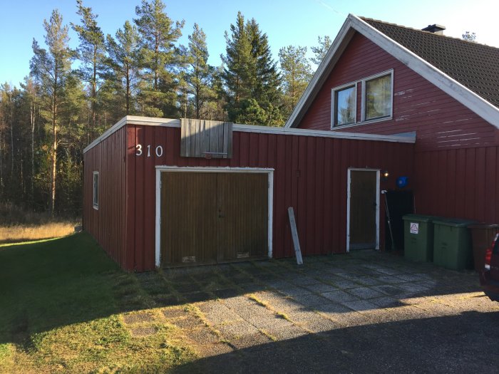 Rödmålat hus med garageport och trädgårdsskjul i solljus, stege lutad mot väggen.