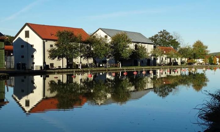 Lugn flod i Söderköping med spegling av vita byggnader och träd under en klarblå himmel.