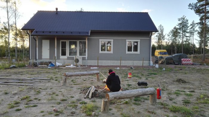 Person sitter vid en eld framför ett pågående husbygge med byggmaterial och verktyg utspritt på marken.