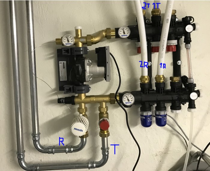 Värmesystem med pump, rör och ventiler på en vägg, några rör märkta med bokstäver.