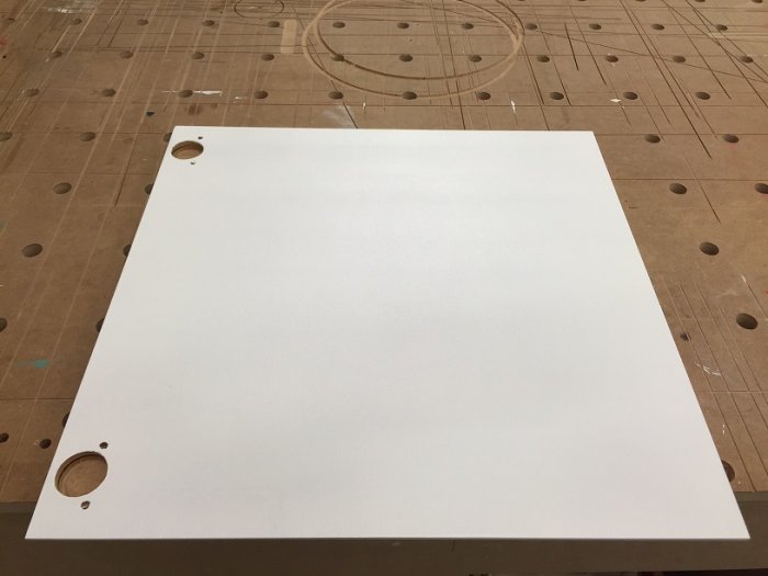 Vitmålad MDF-skåpsfront med hål för handtag, ligger på arbetsbord med hålmönster.