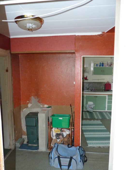 Gammal kamin i hörnet av ett rött rum med synliga murverk, bredvid dörröppning till grönt rum.