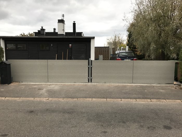 Betongstaket framför svart hus med gånggrind, marksten och aluminiumlister synliga.