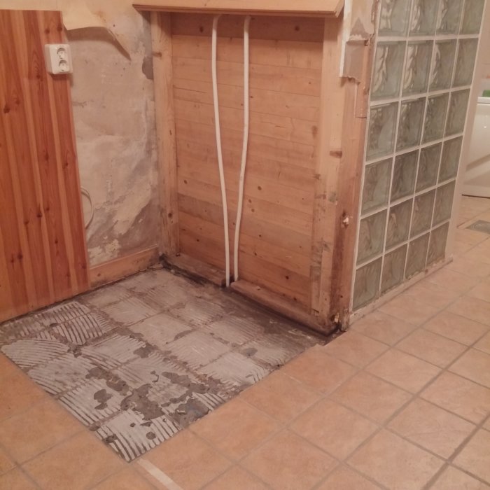 Delvis riven duschhörna med trästomme och skador vid golv och vägg, samt transparent duschvägg.