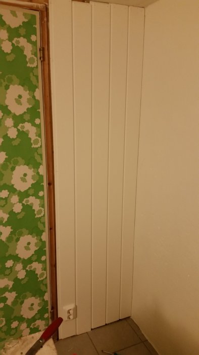 Renovering med nya vita vertikala paneler på vägg bredvid en vägg med grönt blommönstrat tapet.