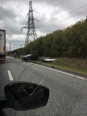 Bil med släpvagn felaktigt lastad står vid vägkanten, med personer och annan trafik i närheten.