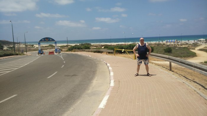 Man i solglasögon och sommarkläder står på en trottoar med sandstrand och hav i bakgrunden.