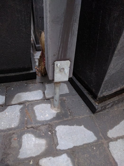 Stolpe med synlig balksko monterad på stenläggning vid svart struktur.