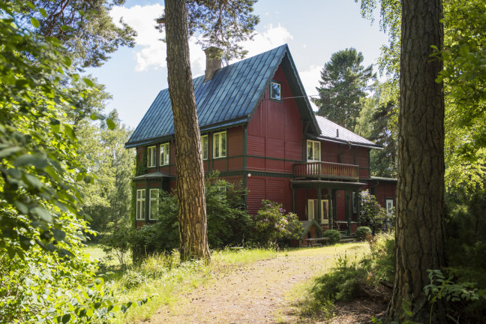 Stor röd villa i äldre stil omgiven av grönska med blått tak och veranda, som representerar 20-tal och äldre arkitektur.