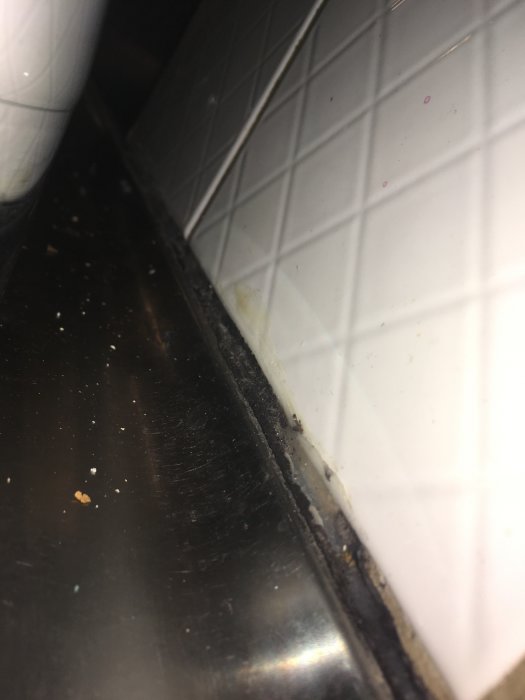 Skador på silikonfog vid diskbänkens kant med svarta klumpar under, mot vita kakelplattor, tecken på mögel eller smuts.