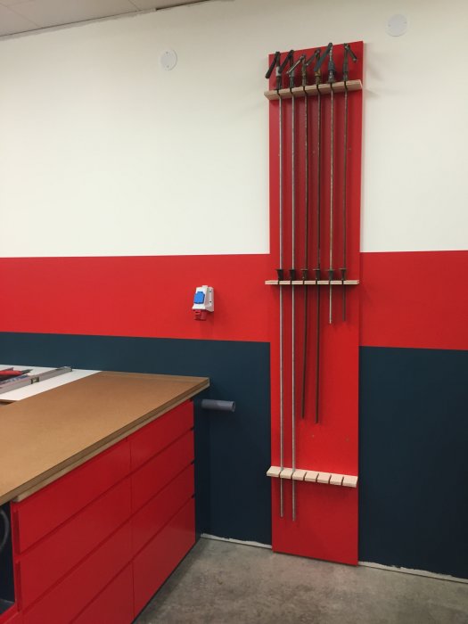 Väggmonterat ställ för limklämmor på en rödmålad och vit vägg ovanför en röd verktygslåda.