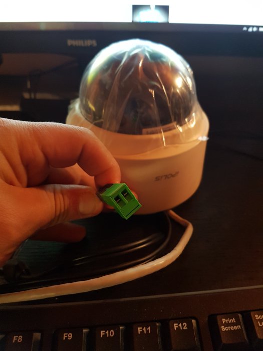 Hand håller en grön strömkontakt framför en övervakningskamera med plastskydd.