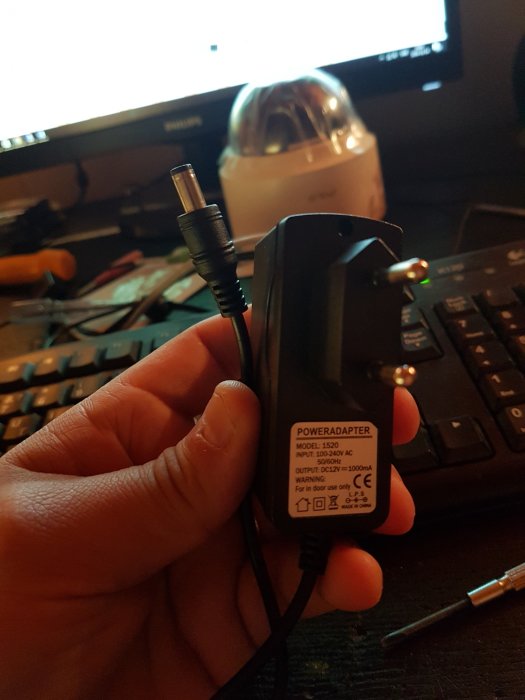 Hand håller en strömadapter med elkontakt och utstickande DC-kontakt framför ett skrivbord.