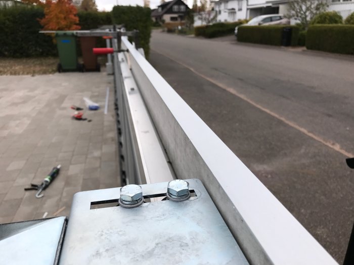 Aluminiumlister monterade på kant med synliga skruvar, suddig bakgrund med verktyg och bostadsområde.