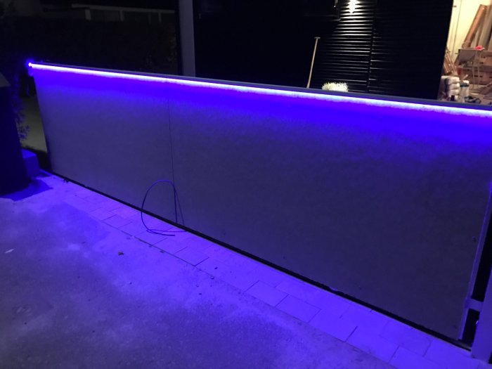 Aluminiumlister med LED-belysningsprofil i blått monterat längs en vägg utomhus på natten.
