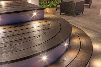 Trädäck i kurva med inbyggda LED-lampor intill ett mörkt bord och en växt. Modern, elegant utomhusdesign.