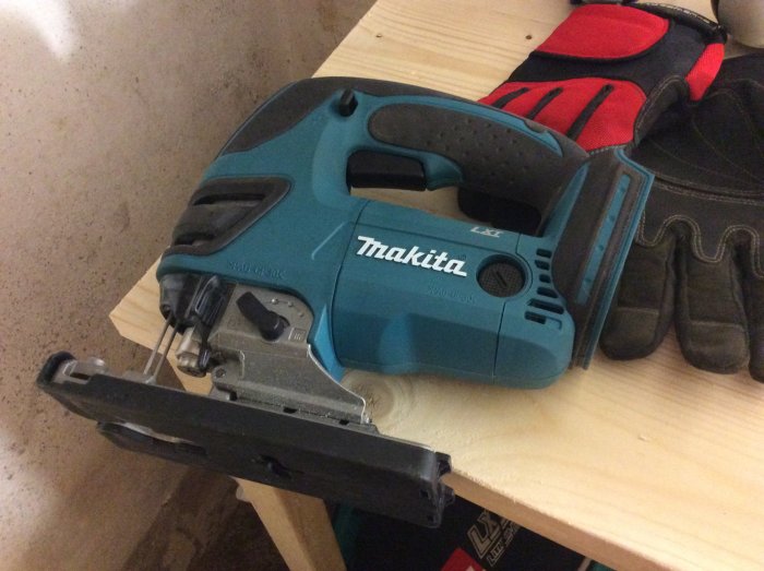 Makita elektrisk sticksåg på träplanka bredvid ett par arbetshandskar.