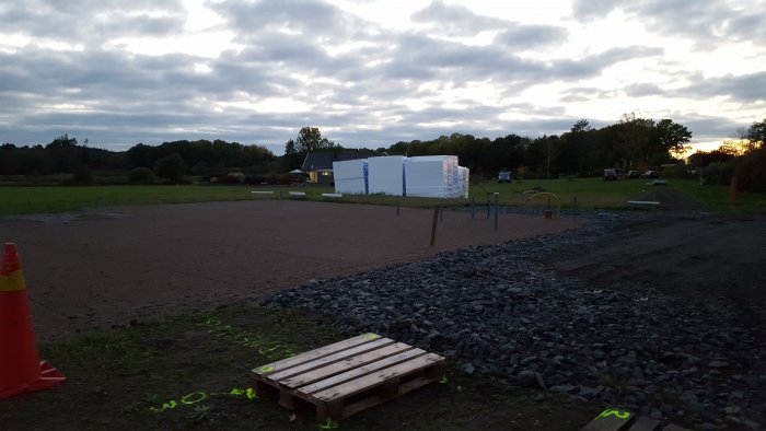 Byggarbetsplats i skymningen med ofixerade rör och isoleringsmaterial på marken.