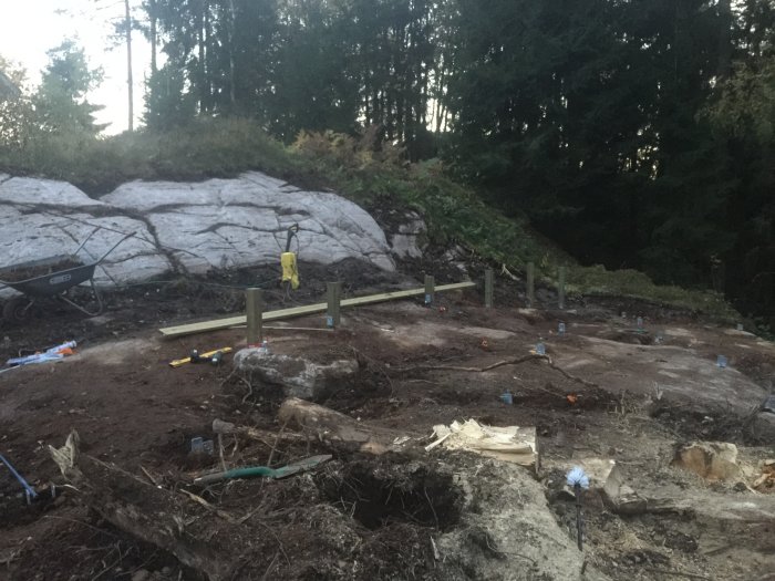Byggplats med stolpar fästa i stolpskor och träreglar som förberedelse för bärlina vid bergskant.