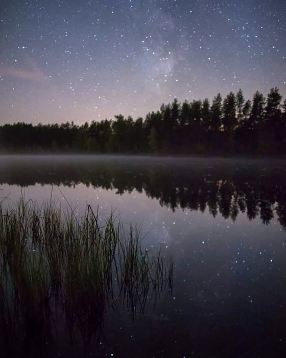 Nattfoto av en sjö med stjärnhimmel, reflektion i vattnet och dimma över ytan.