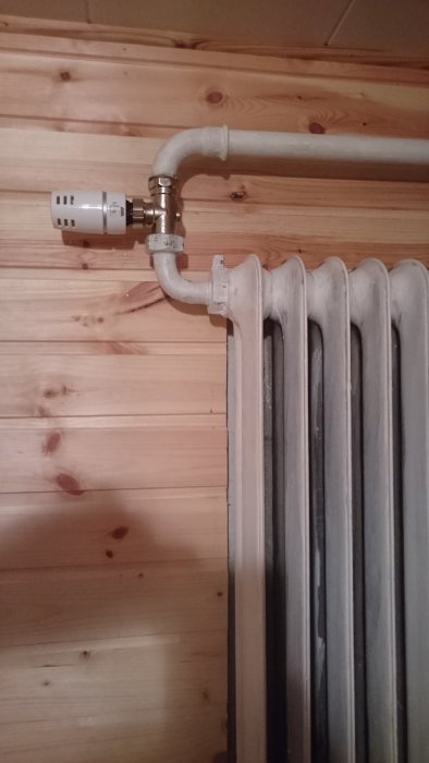 Gammal radiator med ny termostatventil monterad på rörledning mot en trävägg i en källare.