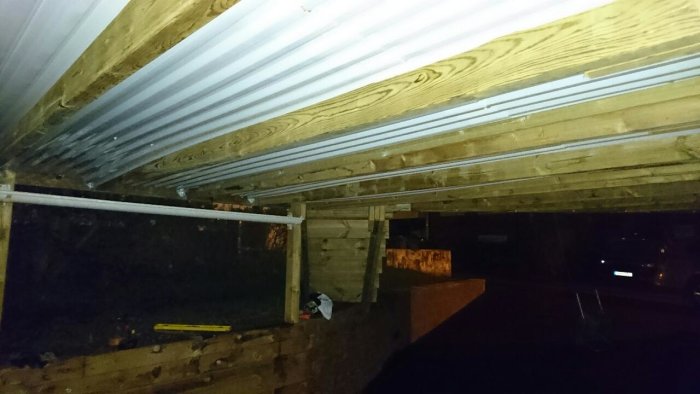 Bild på en enkel carport under en altan med ett provisoriskt plasttak mellan träreglar som läcker vatten.